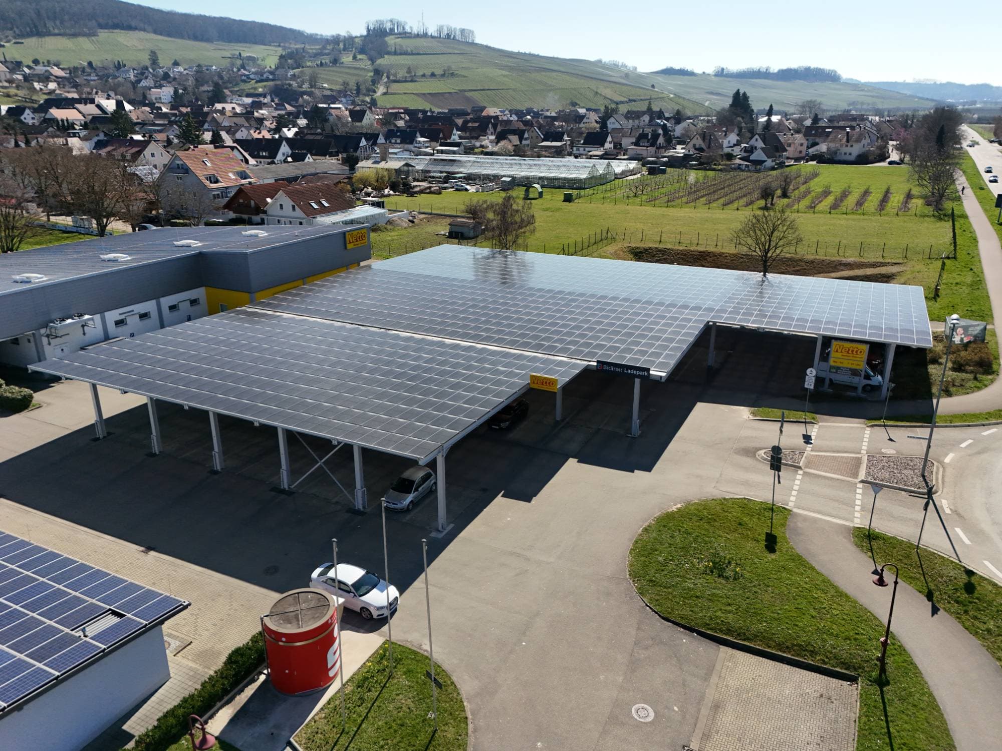 Drohnenaufnahme des Bidirex-Ladeparks Auggen mit PV-Carport neben dem Netto-Markt und Weinbergen im Hintergrund